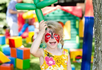 Child with face painting holding a balloon animal in front of bounce house. 