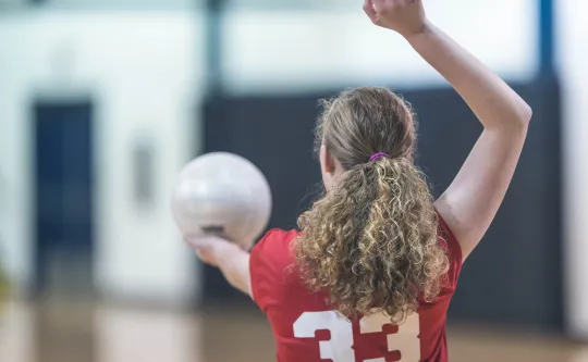 A girl wearing a red jersey raises her hand to serve a volleyball.