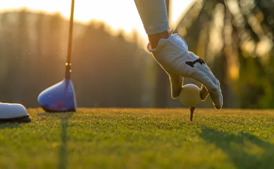 A gloved hand adjusts a golf tee on the golf course. There is a bright sunset and a golf club in the background.