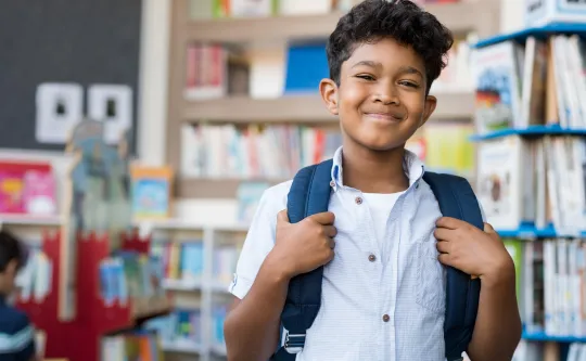 A boy wearing a backpack stands in a classroom and smiles at the camera.