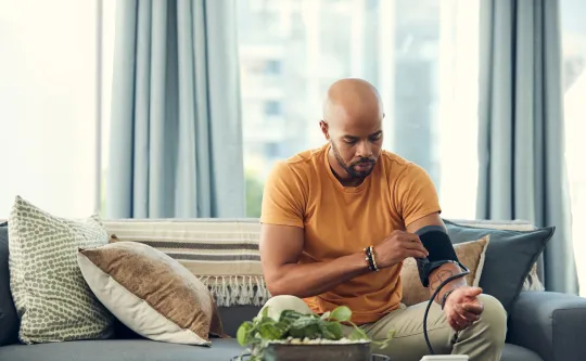 A man monitoring his blood pressure in his home using a blood pressure cuff. He is sitting on a couch.