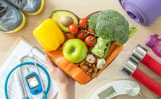 A hand holding a collection of healthy foods. On the table is a collection of fitness equipment, a scale, and other medical devices.