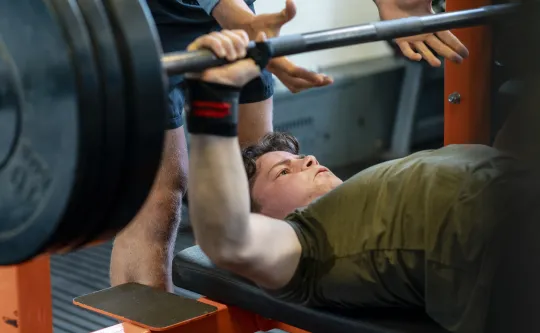 A teenage boy lifting weights while a spotter stands behind him.