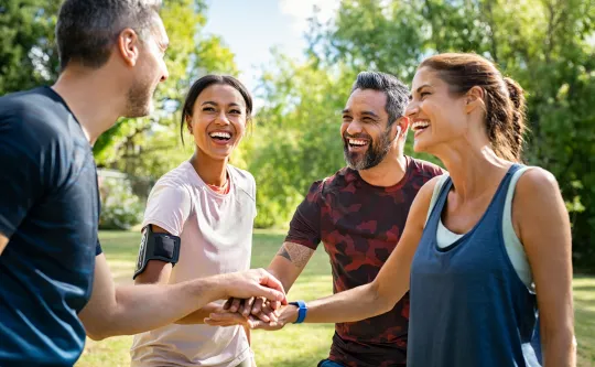 Friends outside standing together in a circle. Their hands are at the center of the circle and they are wearing exercise clothing.