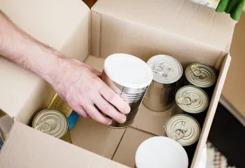 A photo of canned goods inside a cardboard box. There is a man's arm reaching to pick up a can.