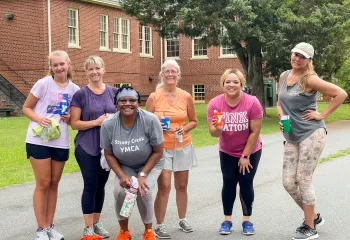 A diverse group of women standing together while smiling at the camera. They are wearing exercise clothing.