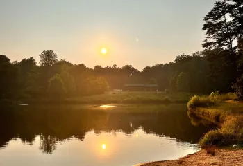 A view overlooking YMCA Camp Weaver's lake. There is a sunset in the background. The sun is warm and yellow and reflected in the lake. In the foreground is a path.