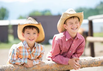 Two young boys dressed in button down shirts and western wear. They are wearing brimmed cowboy hats.