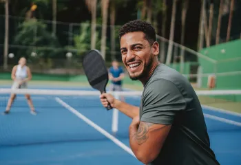 A man playing pickleball outdoors. He is smiling at the camera.