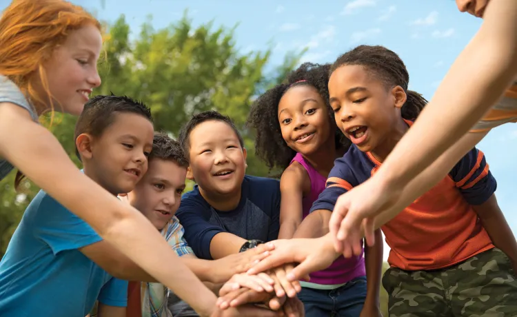 A group of diverse children at summer camp stand in a circle. Their hands are stacked in the middle of the circle.