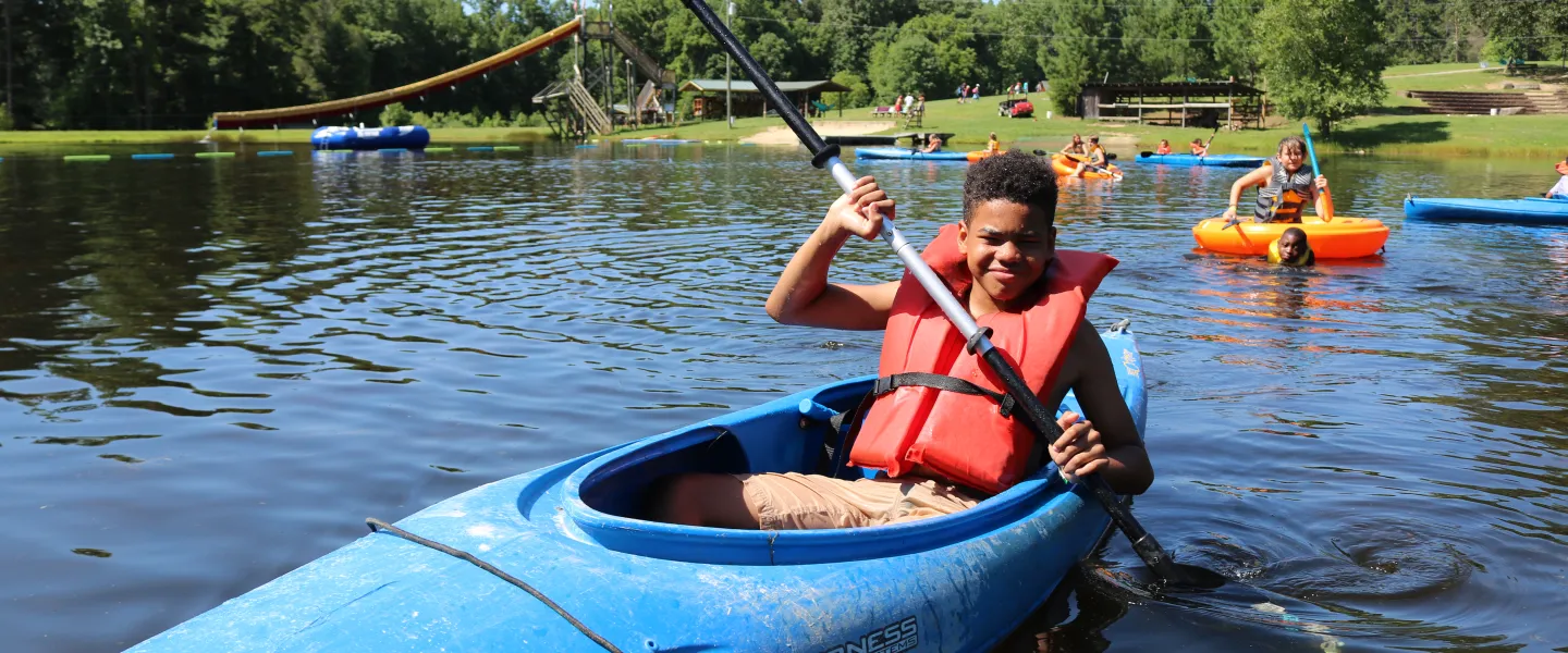 A young boy smiles at the camera while canoeing on a lake.