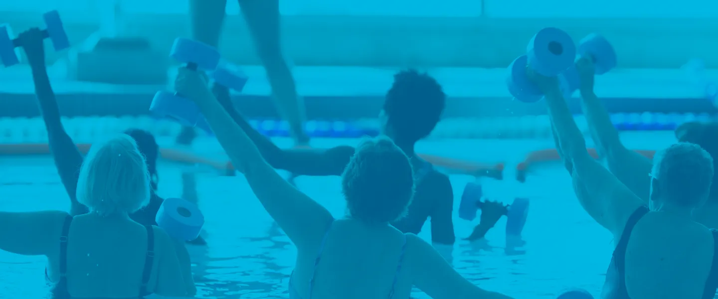 A group of women in a water fitness class. They are exercising with weights and their backs are to the camera while the instructor speaks to them.