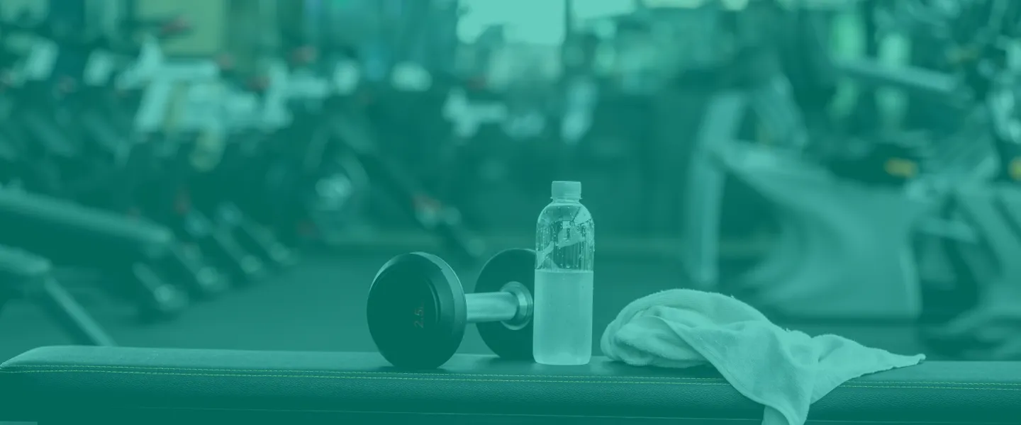 A weight bench in a fitness area. A white towel, a half-filled waterbottle, and a hand weight sit on top of the bench.