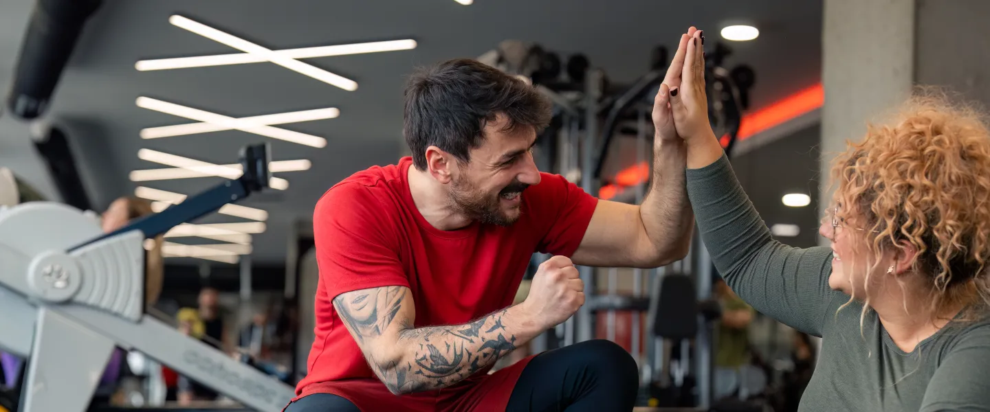 A personal trainer and his client at the gym. The trainer is a man with dark hair and dark facial hair kneeling beside his client, a woman with blond curly hair, sitting on the gym floor. The personal trainer and client are giving each other a high five.