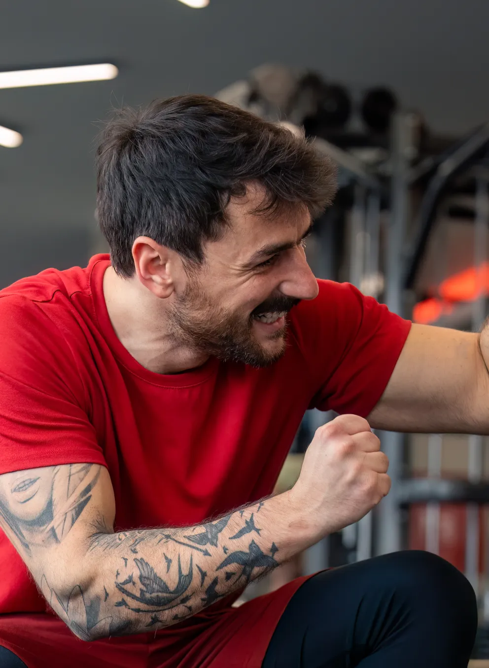 A personal trainer and his client at the gym. The trainer is a man with dark hair and dark facial hair kneeling beside his client, a woman with blond curly hair, sitting on the gym floor. The personal trainer and client are giving each other a high five.