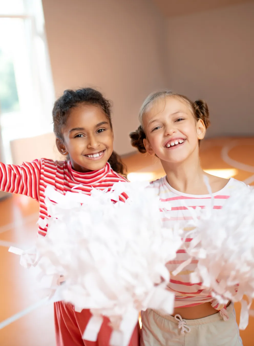 Two young cheerleaders standing together in the gym. They are both holding white cheerleading pom-poms.