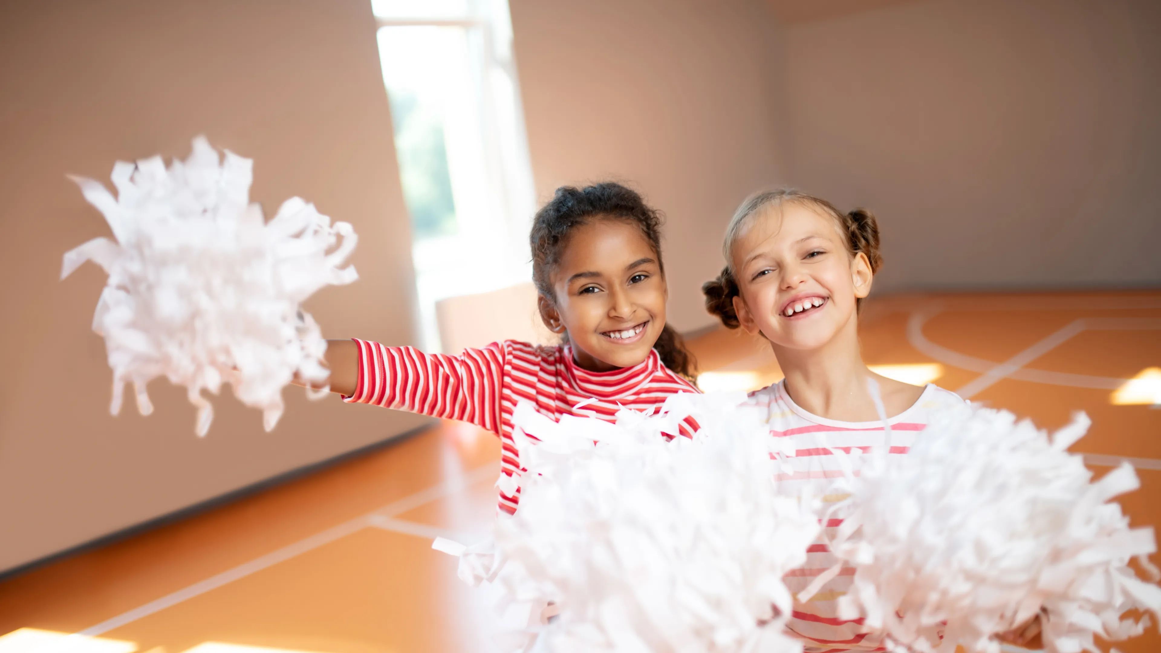 Two young cheerleaders standing together in the gym. They are both holding white cheerleading pom-poms.