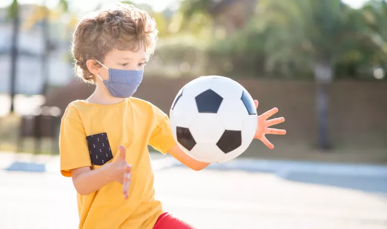 A young boy playing with a soccer ball. He is wearing a mask.