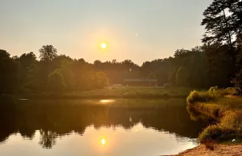 A view overlooking YMCA Camp Weaver's lake. There is a sunset in the background. The sun is warm and yellow and reflected in the lake. In the foreground is a path.