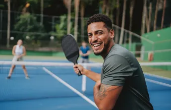 A man playing pickleball outdoors. He is smiling at the camera.