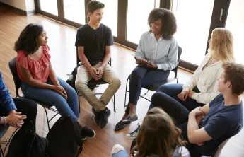 A diverse group of teens sitting together in chairs arranged in a circle. The group is listening to a young woman speak.