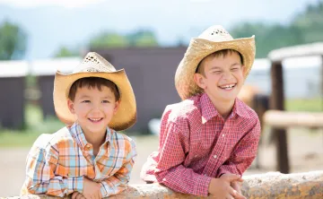 Two young boys dressed in button down shirts and western wear. They are wearing brimmed cowboy hats.