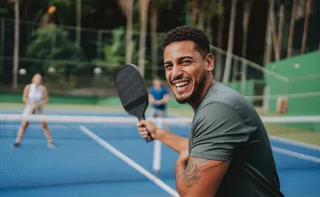 A man playing pickleball outdoors. He is smiling at the camera.